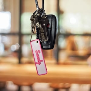 Puede incluir: Un juego de llaves con un llavero de coche negro y un llavero. El llavero es un rectángulo blanco con un borde rosa y el nombre "Sandy" en escritura rosa. Las llaves cuelgan frente a un fondo borroso.