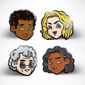 Op de afbeelding: Vier emaille pins met cartoongezichten met verschillende huidtinten, haarkleuren en uitdrukkingen. Elke pin heeft een zwarte omtrek en een glanzende afwerking. De gezichten hebben verschillende oogkleuren en open monden met hoektanden.