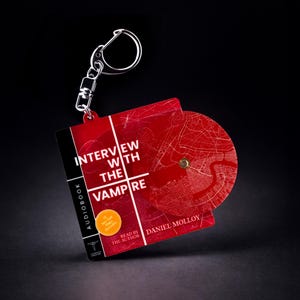Pode incluir: Um porta-chaves quadrado com um design de mapa vermelho e branco com o texto Interview with the Vampire, Audiobook, Daniel Molloy e um autocolante amarelo com #1 New York Times Bestseller. Um disco circular está preso à frente do quadrado.