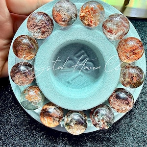 Pode incluir: Uma coleção de contas de cristal redondas e transparentes com inclusões castanho-avermelhadas, dispostas em um padrão circular sobre uma superfície azul clara. As contas são exibidas ao redor de uma depressão central. O texto "Crystal Haven Co." é visível.