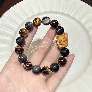 May include: A beaded bracelet featuring a mix of dark, round stones with gold and silver flecks, and a small wooden cat charm. The bracelet is held in a hand, showcasing the natural stone beads.