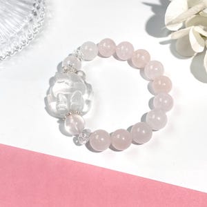 May include: A rose quartz bracelet featuring a carved clear quartz charm and round rose quartz beads. The bracelet has a delicate design with silver accents, perfect for adding a touch of elegance to any outfit.