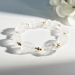 May include: A clear quartz bracelet featuring carved figures and gold-colored beads. The bracelet is strung together with elastic and is displayed on a white surface. The quartz figures are intricately carved and have a frosted appearance.