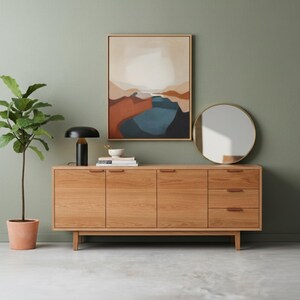 May include: A mid-century modern style wooden sideboard with three doors and three drawers. A black table lamp with a gold base sits on top. A framed abstract landscape painting and a round mirror hang on the sage green wall.