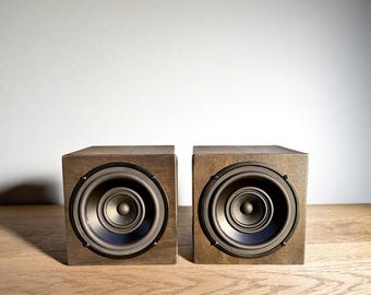 May include: Two wooden speakers with dark brown speaker cones and black trim. The speakers are square-shaped and have a natural wood grain finish. They are positioned side-by-side against a white background, suggesting a stereo pair.