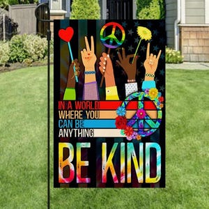 Può includere: Una bandiera da giardino con sfondo nero con la scritta "In a world where you can be anything, be kind". La bandiera presenta un simbolo di pace, mani e fiori in un design colorato. Un cuore e un fiore sono sollevati.