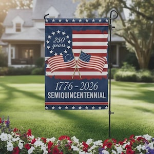 Op de afbeelding: Een patriottische tuinvaandel met het ontwerp van de Amerikaanse vlag en de tekst "250 years" en "1776-2026 Semiquincentennial". De vlag, op een zwarte metalen standaard, toont twee gekruiste vlaggen in een tuin.