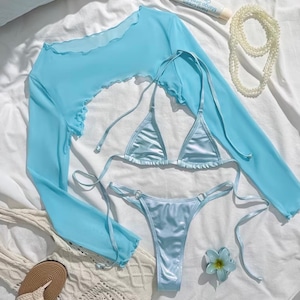 Könnte beinhalten: Hellblaues Bikini-Set mit langärmligem Oberteil, String-Hose und Triangel-Top. Das Set enthält ein passendes transparentes Oberteil mit Rüschenkanten. Eine weiße Perlenkette, Sandalen und eine cremefarbene Tasche sind ebenfalls im Bild.