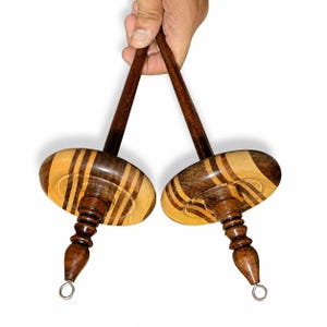 Può includere: Due fusi in legno scuro e chiaro, caratterizzati da una fusaiola circolare con motivi a strisce alternate, un lungo stelo affusolato, un manico tornito decorativo e un piccolo gancio metallico sulla punta inferiore per la filatura.