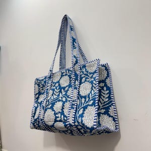 May include: A blue tote bag with a white floral pattern. The bag has a quilted design with white and blue striped trim and handles. The bag is hanging on a white wall.
