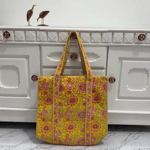 May include: A yellow tote bag with a floral pattern in pink and gray. The bag has long handles and is made of quilted fabric. The background includes a white cabinet with decorative knobs and two bird figurines.