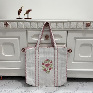 May include: White tote bag with red trim and handles. The bag features a floral design in the center. The bag is made of a quilted material. The background includes a white cabinet with decorative knobs and two bird figurines.