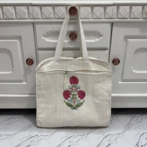 May include: Cream-colored quilted tote bag with a floral embroidery design in shades of pink, green, and blue. The bag has a zipper closure and a handle for carrying. The tote bag is hanging on a white cabinet.