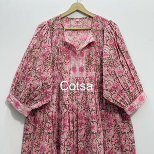 Puede incluir: Vestido rosa con estampado floral, mangas tres cuartos y cuello redondo. El vestido presenta un delicado estampado floral en tonos rosas y verdes, con la palabra "Cotsa" impresa en la parte delantera. El vestido está colgado de una percha de madera.