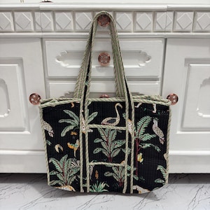 May include: Black tote bag with a tropical bird and plant pattern. The bag has a striped green and white trim and handles. The bag is sitting in front of a white cabinet with rose gold knobs.