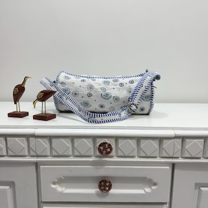 May include: A white and blue patterned shoulder bag with a matching strap. The bag features a cylindrical shape and a repeating eye-like design. Two decorative bird figurines are on the left.