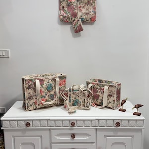 May include: A collection of floral-patterned bags in various sizes, including a tote, a handbag, and a small pouch. The bags feature a cream background with red, blue, and green floral designs. The bags are displayed on a white cabinet.