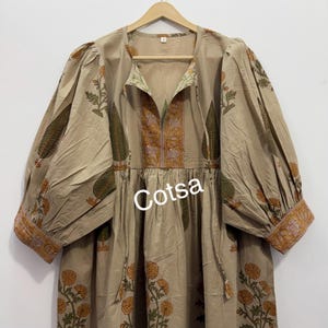 Peut inclure: Robe beige ornée de motifs floraux et de feuilles verts et oranges. La robe présente de longues manches bouffantes et une encolure en V avec des bordures décoratives. Le mot "Cotsa" est imprimé sur le devant de la robe.