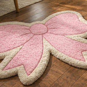 Può includere: Un tappeto rosa a forma di fiocco con bordo color crema su un pavimento in legno. Il centro e i dettagli del fiocco sono rosa strutturato, con un bordo color crema morbido. Questo tappeto aggiunge un tocco decorativo a qualsiasi stanza.
