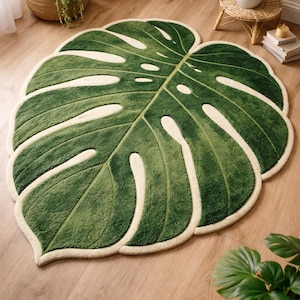 Puede incluir: Una alfombra grande con mechones en forma de hoja de Monstera. La alfombra presenta un color verde bosque profundo con detalles en color crema que resaltan las venas y las fenestraciones características de la hoja, creando un diseño botánico detallado.
