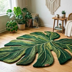 May include: A large, tufted area rug shaped like a monstera leaf, featuring a gradient of deep forest green, olive, and lime green tones with intricate, raised textures that mimic the natural veins and lobes of a tropical plant leaf.