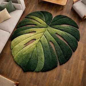 Op de afbeelding: Een groene, Monstera-blad-vormige vloerkleed met een getextureerd oppervlak en uitgesneden details. Het ontwerp van het vloerkleed bevat verschillende groentinten, waardoor een driedimensionaal effect ontstaat. Het ligt op een houten vloer, met een bank en stoelen op de achtergrond.
