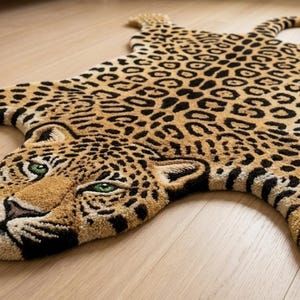 May include: A tufted area rug shaped like a leopard, featuring a tan and cream base with black and brown spots. The leopard head has detailed facial features, including green eyes, a black nose, and whiskers, with four extended paws and a tail.