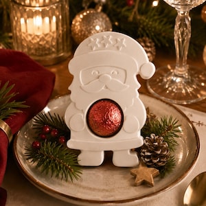 Könnte beinhalten: Eine weiße Dekofigur in Form eines Weihnachtsmanns mit glatter, matter Oberfläche und einer kreisförmigen Aussparung in der Mitte, in der eine runde, in glänzende rote Folie eingewickelte Schokoladenkugel liegt. Die Figur trägt eine Mütze mit drei Sternen.