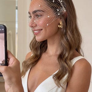 May include: A white bridal veil with a net design and scattered pearls. The veil is attached to a white headband. The woman is wearing gold flower earrings and a white dress. The woman is holding a phone.