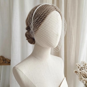 May include: A white bridal veil with a delicate net design is displayed on a mannequin. The veil is attached to a thin headband. The mannequin is wearing a white strapless dress, and the background includes white curtains and a mirror.