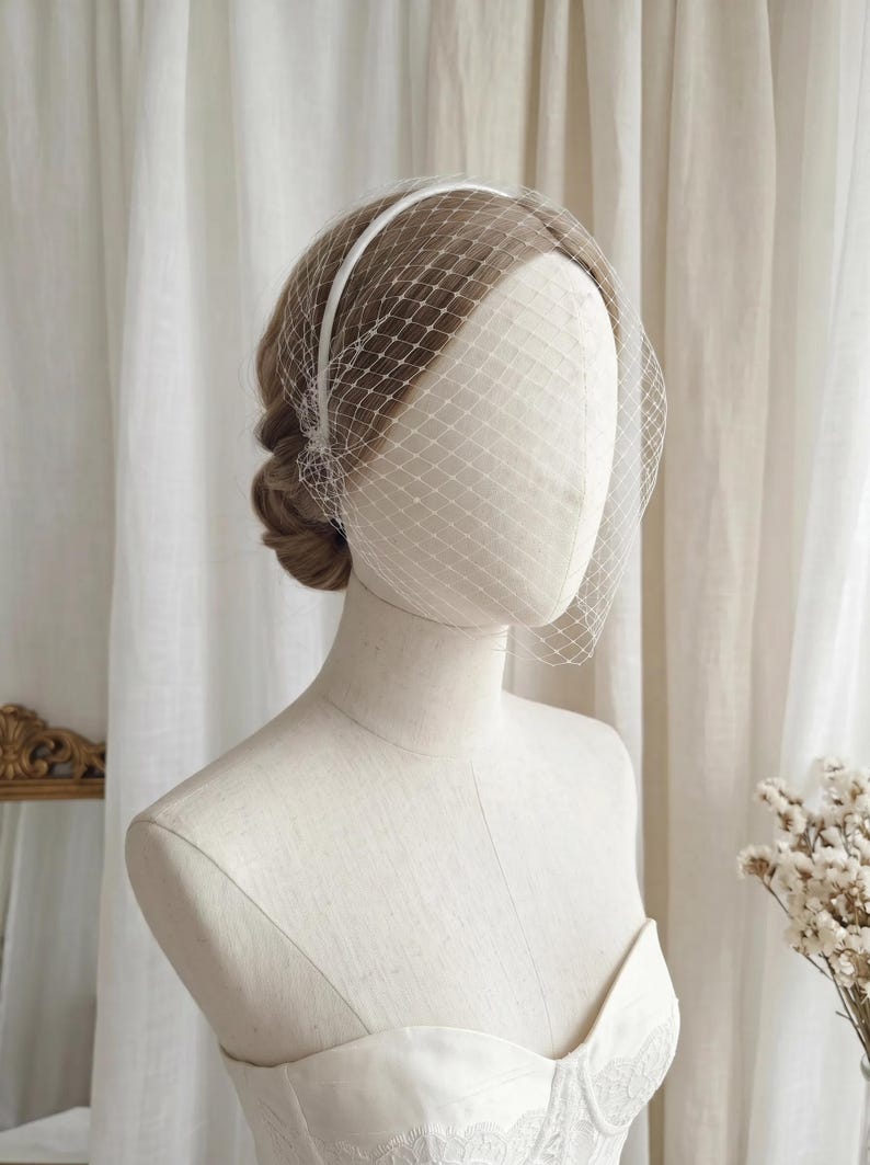 May include: A white bridal veil with a delicate net design is displayed on a mannequin. The veil is attached to a thin headband. The mannequin is wearing a white strapless dress, and the background includes white curtains and a mirror.