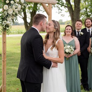 May include: A wedding ceremony with a bride and groom kissing under a wooden arch decorated with white flowers. The bride wears a white dress and veil, while the groom wears a black suit. Bridesmaids in green dresses and groomsmen in black suits stand in the background.