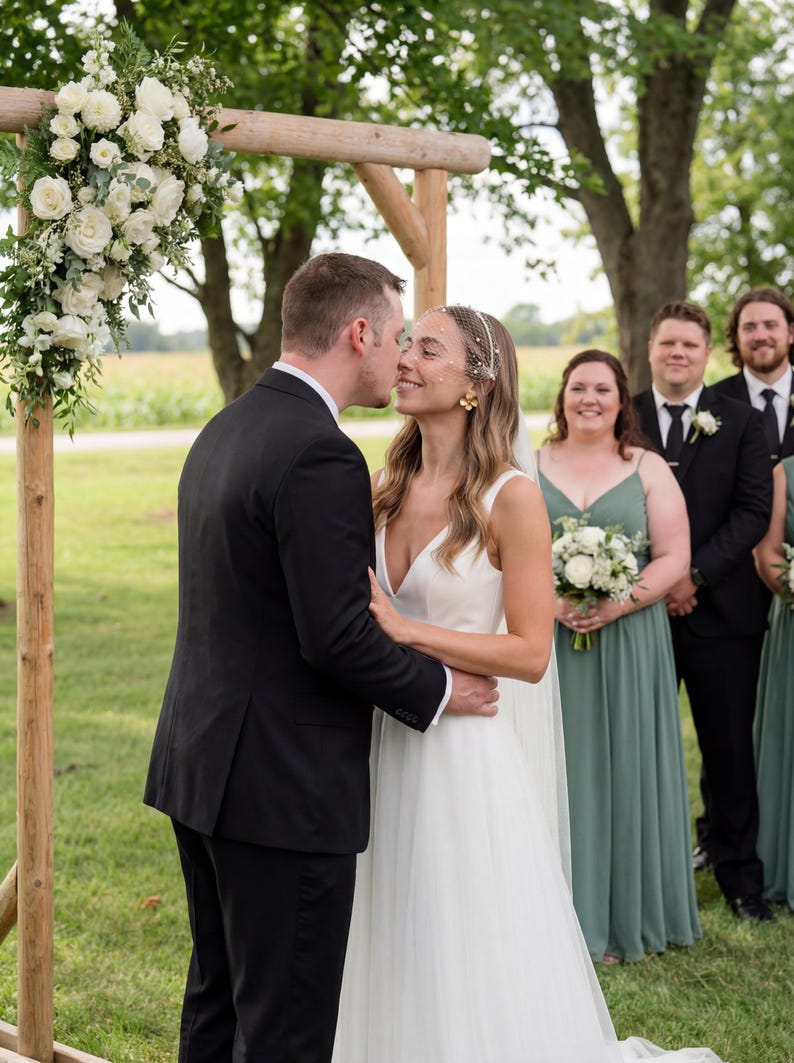 May include: A wedding ceremony with a bride and groom kissing under a wooden arch decorated with white flowers. The bride wears a white dress and veil, while the groom wears a black suit. Bridesmaids in green dresses and groomsmen in black suits stand in the background.