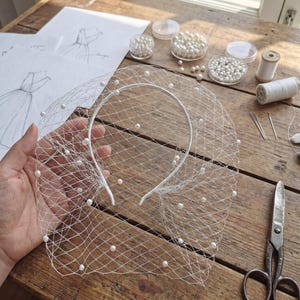 May include: A white bridal veil with scattered pearls, held over a wooden surface. The veil is attached to a white headband. Other items on the table include sketches, pearls, thread, and scissors, suggesting a craft or design project.
