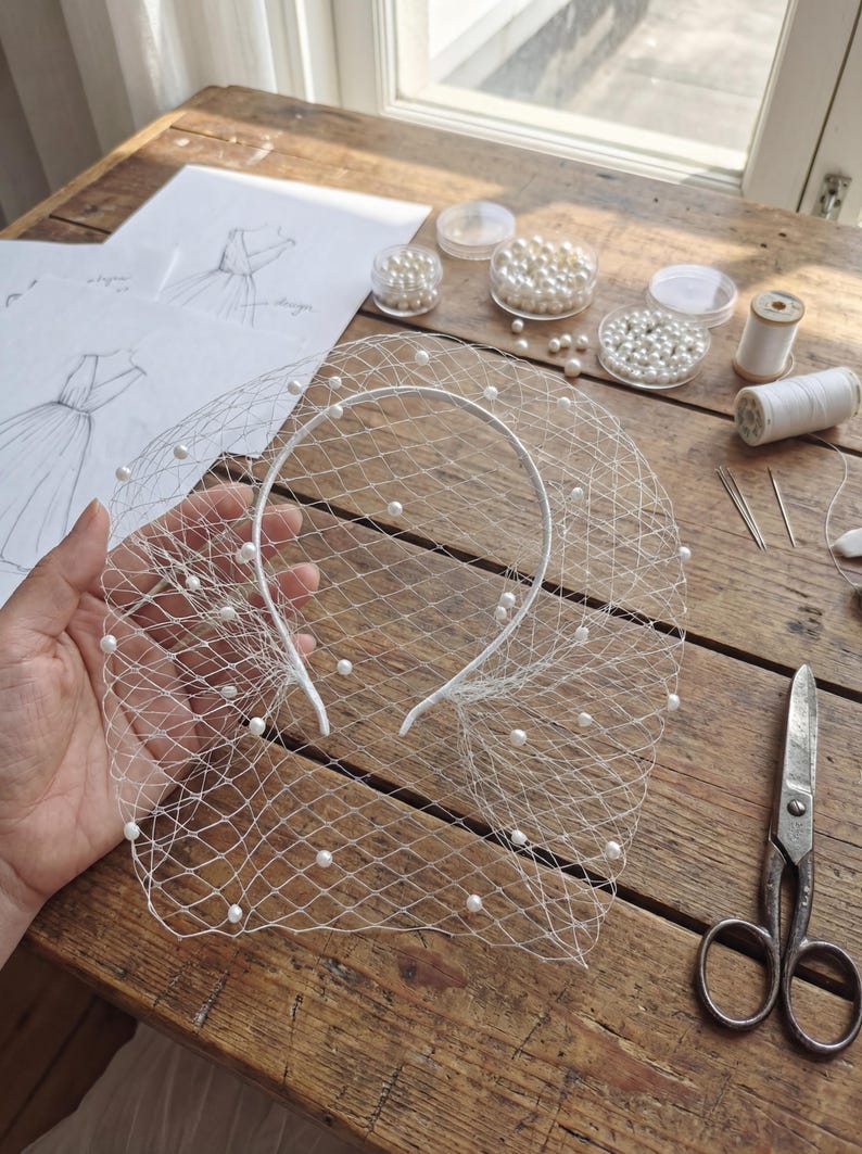 May include: A white bridal veil with scattered pearls, held over a wooden surface. The veil is attached to a white headband. Other items on the table include sketches, pearls, thread, and scissors, suggesting a craft or design project.