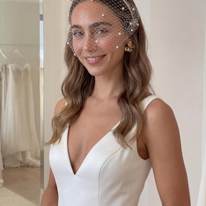 May include: A bride wearing a white dress and a pearl-studded veil. The veil is attached to a white headband and drapes over the face. Gold flower earrings and a bracelet complete the look. The background includes a mirror and white dresses.