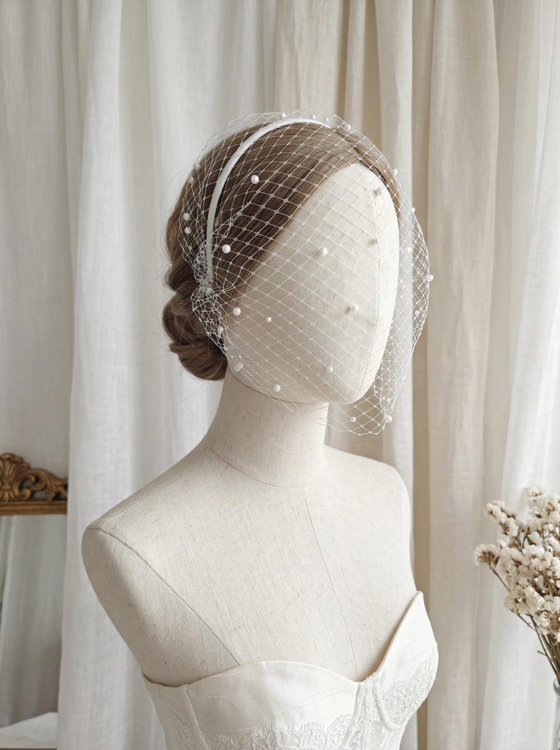 May include: A bridal headpiece featuring a white headband and a delicate net veil. The veil is adorned with scattered, small, white pearls. The headpiece is displayed on a mannequin, showcasing its design and details. The overall aesthetic is elegant and suitable for weddings.