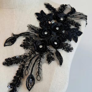 May include: Black floral lace applique with beaded and sequined accents. The applique features three-dimensional fabric flowers with rhinestone centers, and delicate leaf and feather details. The design is mounted on a sheer mesh backing.