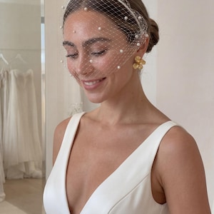 May include: A bride wearing a white wedding dress with a deep V-neckline. She is wearing a delicate white veil with pearl accents and gold flower earrings. The background shows a bridal shop with wedding dresses.