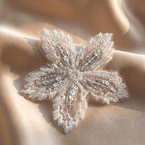 May include: A detailed, five-petal floral applique. The applique is made of off-white fabric, embellished with sequins and beads. The flower is set against a satin background in a warm, neutral tone.