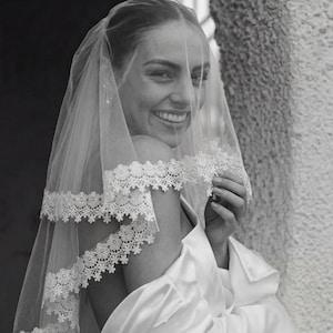 Könnte beinhalten: Eine lächelnde Person trägt ein weißes Hochzeitskleid und einen transparenten Schleier mit Spitzenbesatz. Der Schleier ist mehrschichtig und fällt über die Schultern. Das Bild ist in Schwarzweiß.