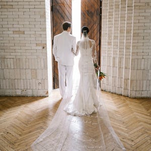 Puede incluir: Una pareja camina por una puerta, la novia con un vestido de novia blanco con un velo largo. El velo tiene una escritura impresa. El novio lleva un traje blanco. El suelo es de madera.