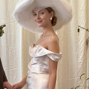 Puede incluir: Una mujer con un vestido blanco con hombros descubiertos y un sombrero grande de tul. También lleva pendientes de perlas. El vestido tiene un brillo metálico y el sombrero es voluminoso. El fondo es una cortina color crema.