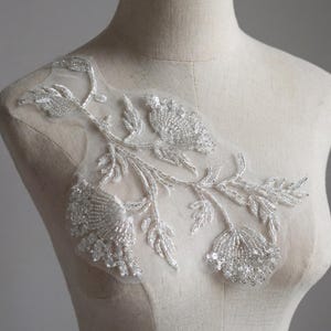 May include: A detailed floral applique with beaded flowers and leaves in white and silver. The applique is attached to a sheer, off-white mesh backing. This decorative embellishment is suitable for bridal wear or other special occasion garments.