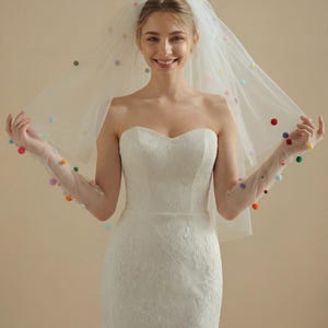 Puede incluir: Vestido de novia blanco de encaje sin tirantes con escote corazón. La novia lleva un velo transparente y guantes adornados con pompones de colores. El velo está drapeado sobre la cabeza y los hombros.