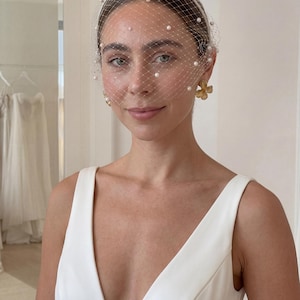 May include: A bride wearing a white veil with scattered pearls and a matching headband. The veil is a fine net that covers the face. She is wearing gold flower earrings and a white sleeveless dress with a deep V-neck.