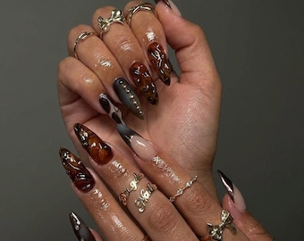 May include: A hand displaying long, almond-shaped acrylic nails in various designs. Some nails are a glossy brown with a marbled effect, others are a gradient of black and nude, and some feature gold accents. Gold rings adorn the fingers.