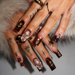 Może przedstawiać: Długie, kwadratowe sztuczne paznokcie z ciemnobrązowym wzorem szylkretowym, francuskim manicure i trójwymiarowymi zdobieniami kwiatowymi. Wzór zawiera małe złote zawieszki, przezroczyste cyrkonie i motyw wiśni. Widoczny jest napis @Icynails4u.
