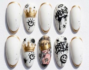 May include: A set of ten almond-shaped press-on nails featuring a white base with gold tips. Designs include raised gold crowns, black graffiti-style text reading MADRID and PSYCH, a cartoon character wearing a gold crown, and black splatter accents.