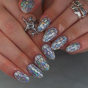 May include: Close-up of a hand with iridescent, geometric nail art. The nails have a clear base with a mosaic-like pattern that reflects multiple colours. A silver ring with a starburst design and a ring with the word "forever" are also visible.